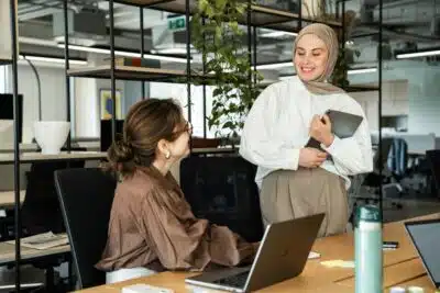 Two women in an office talking. One is seated by a laptop, talking to a woman in a hijab who is standing next to her. By Ahmed on Pexels