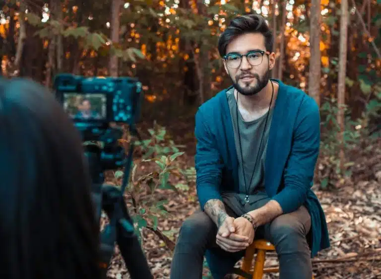 A young man sits in front of a camera outside by some trees. By Rodrigo Souza on pexels