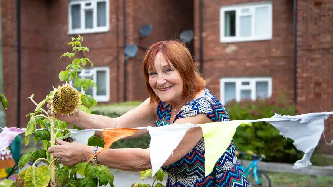 A woman outside some flats holding a sunflower and putting up some bunting to celebrate.